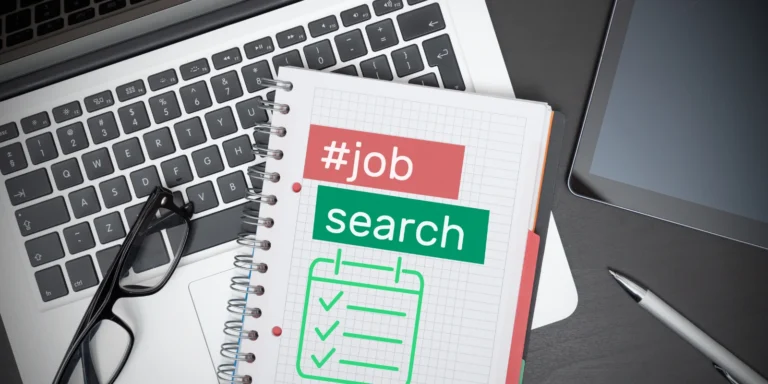 A close-up of a notebook with a job search label, checkboxes, and a pen on a desk, symbolizing the job search checklist with a laptop and tablet in the background.